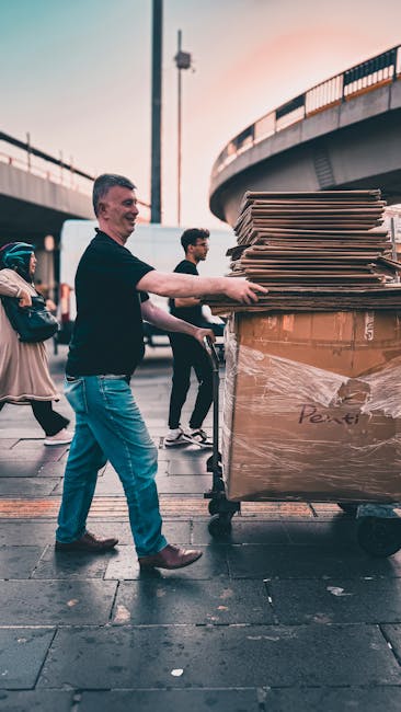 A man is pushing a large wheelie bin filled with flattened cardboard boxes and packaging materials under an overpass or raised road structure during dusk, with a group of pedestrians walking nearby. The bin is made of dark plastic and has a substantial, sturdy appearance. The flattened cardboard is light brown with some visible printed labels. The scene is outdoors on a paved surface, with the bridge or elevated roadway curving in the background, and a tall streetlamp standing to the left of the man. The ambient lighting suggests late afternoon or early evening, with the sky showing soft, warm tones. The presence of the bin and scattered cardboard indicates a context of waste collection or rubbish removal, representing private waste disposal solutions, such as those offered by rubbish removal services like Rubbish Removal Elephant and Castle. The overall environment appears urban, with other pedestrians and city infrastructure subtly visible in the background, emphasizing a professional and observational tone suitable for service-related content.