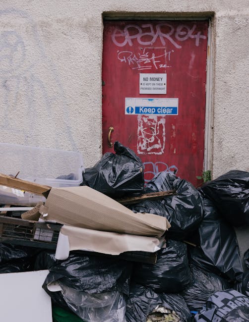 A pile of black rubbish bags filled with waste materials is stacked against a beige, textured wall next to a red metal door that is covered in graffiti and stickers. The bags are tightly sealed, with some appearing bulging from the contents inside, and are placed on a concrete surface, possibly a pavement or alleyway. Among the rubbish, a partially collapsed cardboard box and a piece of undelivered white packaging material are visible, along with some loose paper and small debris. The red door features a small rectangular sign indicating 'No Money' and a larger blue and white sticker with the text 'Keep clear'. A faintly visible green plant is near the base of the wall beside the rubbish. The scene suggests an informal rubbish accumulation, possibly related to private or alternative waste collection methods, and is typical of an area where independent rubbish removal services, like those offered by rubbishremovalelephantandcastle.co.uk, might be involved in clearing discarded waste from urban or residential environments. The lighting is neutral, with no strong shadows or highlights, conveying a straightforward, observational vibe aligned with waste management contexts.