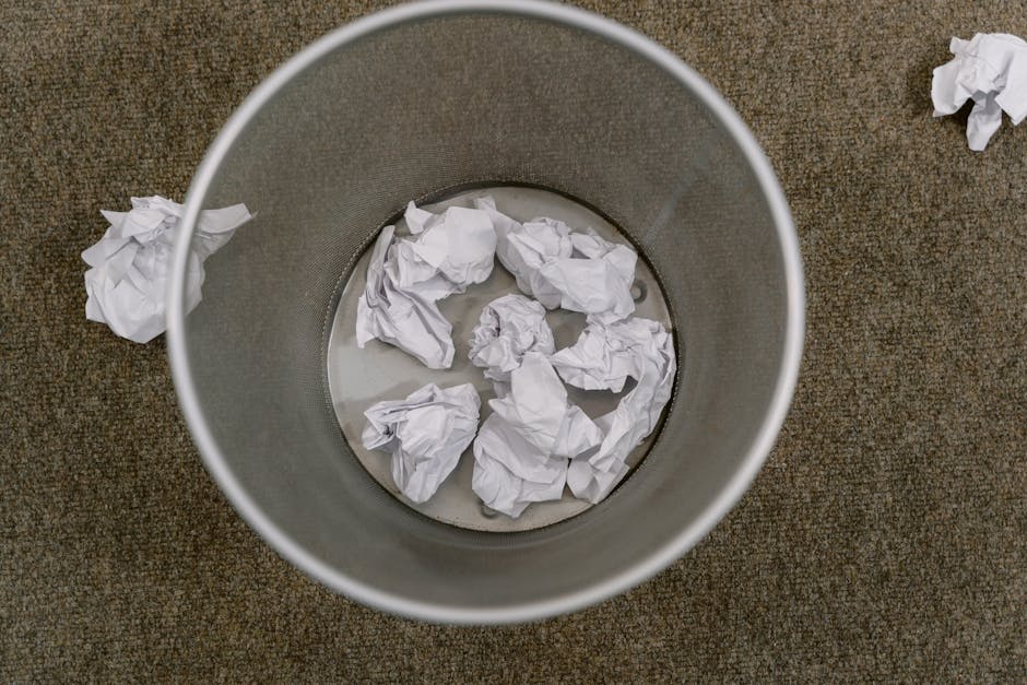 A cylindrical metal waste bin with a matte silver finish, placed on a brown textured carpeted floor. The bin contains several crumpled sheets of white paper, some submerged in a small amount of water at the bottom of the bin. Outside the bin, a few additional crumpled paper pieces are scattered on the carpet. The environmental setting appears to be an indoor office space or similar interior environment, with the focus on waste collection and paper disposal. The scene illustrates a typical private rubbish collection scenario involving uncollected paper waste in a home or office setting, aligned with alternative waste handling services such as independent rubbish removal.