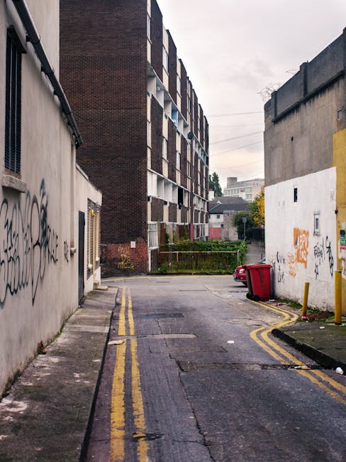 A narrow urban alleyway or backstreet situated between two weathered buildings. On the left, a plain concrete wall with black graffiti tags, and on the right, a similar white wall also covered in graffiti and street art. The ground is paved with asphalt, showing signs of wear and patches, with two narrow yellow lines painted along the edges indicating no-parking zones. A red wheelie bin is tipped over against the white wall, near a small yellow bollard, and a faint puddle reflects the dull, overcast sky overhead. In the background, a larger multi-storey brick residential building with a mix of small balcony windows and air conditioning units is visible, contrasting with the more utilitarian, graffiti-marked surroundings. The environment suggests an area where private waste collection or rubbish removal services such as those provided by Rubbish Clearance Elephant and Castle may operate, with the scene capturing typical urban waste disposal points in a densely built-up locality.