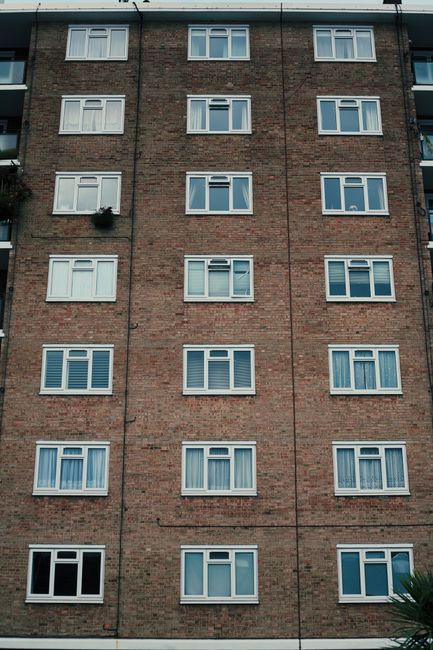 A tall multi-storey brick residential building featuring five levels of windows with white frames, arranged in a regular grid pattern. Each level has a consistent layout with the windows appearing to be double-glazed, some with curtains or blinds partially visible behind the glass. The brick facade is reddish-brown with a slightly weathered texture, and the building's corner and edges are clearly defined. The lower left window on the ground floor is open, displaying a black interior, while the other windows are closed. In the upper left corner of the image, a small potted plant with dark green foliage extends from a window box. The sky above the building is not visible, but the diffuse natural lighting suggests an overcast day. The scene emphasizes the uniformity and straightforward construction typical of residential blocks, with no visible rubbish or waste management activities included in the visual. This detailed exterior shot may indirectly support discussions around urban living and the importance of responsible waste disposal, as seen in associated rubbish removal services.