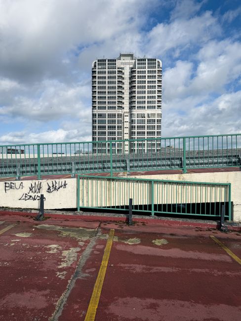 A narrow urban alleyway or backstreet situated between two weathered buildings. On the left, a plain concrete wall with black graffiti tags, and on the right, a similar white wall also covered in graffiti and street art. The ground is paved with asphalt, showing signs of wear and patches, with two narrow yellow lines painted along the edges indicating no-parking zones. A red wheelie bin is tipped over against the white wall, near a small yellow bollard, and a faint puddle reflects the dull, overcast sky overhead. In the background, a larger multi-storey brick residential building with a mix of small balcony windows and air conditioning units is visible, contrasting with the more utilitarian, graffiti-marked surroundings. The environment suggests an area where private waste collection or rubbish removal services such as those provided by Rubbish Clearance Elephant and Castle may operate, with the scene capturing typical urban waste disposal points in a densely built-up locality.