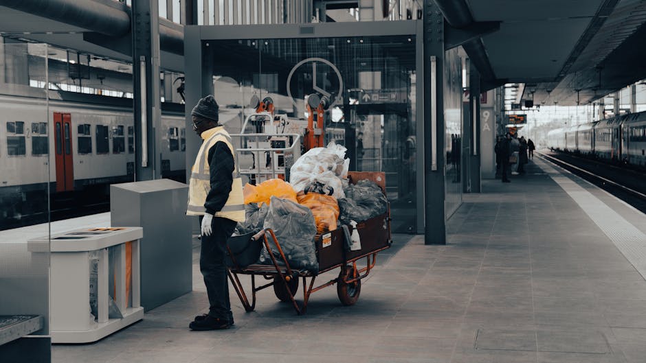 A man wearing a dark jacket, grey beanie, and reflective safety vest stands on a modern train station platform next to a large red wheelbarrow filled with various types of waste, including black, grey, and orange trash bags, as well as some loose debris. The platform features smooth, grey stone paving and is sheltered by a contemporary metal and glass canopy. Behind the man, there is a glass-enclosed lift or elevator, and in the background, a distant train with closed doors is visible along with a few other platform users walking or waiting. The overall scene appears to depict a private or alternative waste collection process, with the man possibly involved in rubbish removal or clearance services, supporting the logistical needs of waste disposal at the station environment. The lighting suggests natural daylight, and the surroundings are clean and orderly, consistent with urban infrastructure focused on efficient transport and waste management.