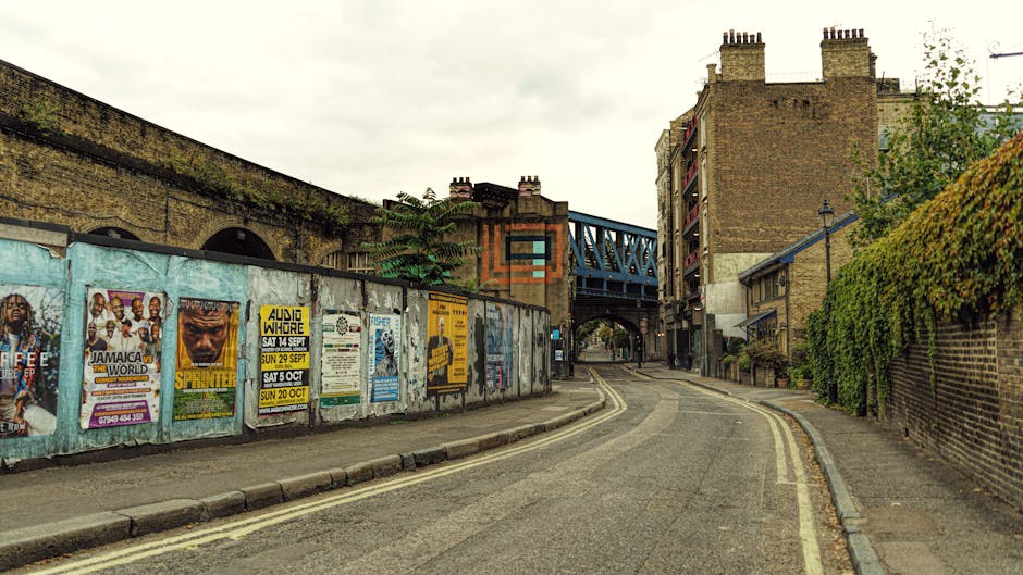 A narrow urban street scene featuring a curved asphalt road with double yellow lines on the side, flanked by brick buildings on the right and a tall brick wall on the left decorated with multiple colorful posters and flyers. The posters display various event advertisements, including music shows and promotional messages, with some partially peeling or weathered. Behind the wall, there is a blue metal bridge structure extending over the street, connecting the buildings, and a large, aged brick building with multiple chimneys dominates the background under an overcast sky. The right side of the street is lined with a green hedge climbing a brick wall, while the sidewalk appears cracked and uneven. The environment suggests an inner-city area undergoing maintenance or private waste handling, with the scene implying an alternative to municipal rubbish collection, potentially associated with local rubbish removal services such as those offered by Rubbish Removal Elephant and Castle, situated near Elephant and Castle station.
