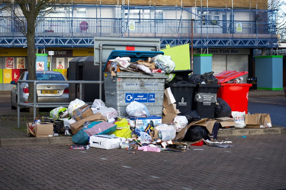 An outdoor urban scene showing a large collection of overflowing rubbish bags and scattered waste material piled around multiple public recycling bins and a grey rubbish dumpster with a partially open lid, situated on a paved area adjacent to a sidewalk. The waste includes various black and green plastic rubbish bags, cardboard boxes, crumpled paper, plastic packaging, and other packaging debris. The background features a building with a yellow storefront, signage, and a blue metal scaffolding structure covering the upper part of the facade. A parked silver car is visible on the left side of the image, with a tree behind the parked vehicle. The scene appears to reflect a private waste collection or rubbish removal service in progress, with a range of different waste materials prepared for disposal or recycling, situated in an environment typical of urban waste management areas.