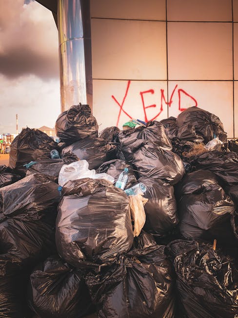 A large collection of black rubbish bags, some appearing filled with waste materials, positioned against a tiled wall on an urban pavement. Several clear plastic bottles are visible on or near the bags, indicating typical household waste. The bags are stacked and leaning slightly, with a few partially torn or crumpled. Behind the bags, on the tiled wall, there is red graffiti spelling out the word 'XEND' which suggests an informal or unauthorized disposal site. The surrounding environment includes a cloudy sky and distant cityscape visible to the left, with warm lighting that highlights the textures of the plastic bags and the reflective surface of a standing metallic structure partially visible on the left. The scene suggests an instance of private waste accumulation, potentially due to access issues or alternative rubbish collection arrangements. The overall setting emphasizes an urban area where rubbish removal services like those offered by rubbishremovalelephantandcastle.co.uk may be needed to manage unmanaged waste like this.