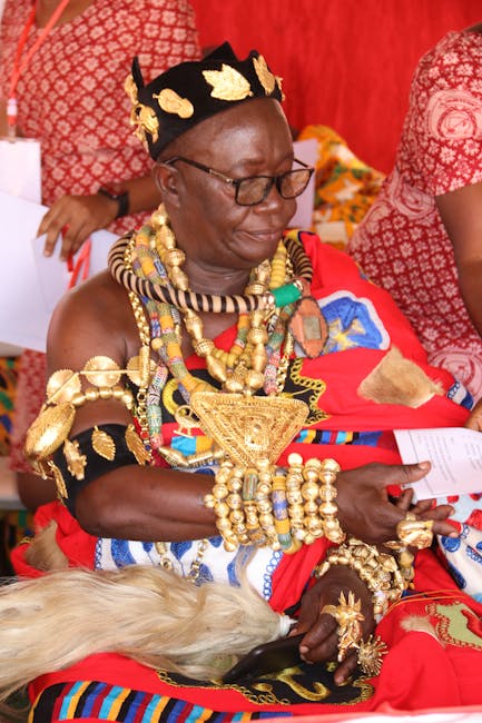 A woman wearing traditional African attire and elaborate gold jewelry, including multiple necklaces, bracelets, and a large triangular pendant, seated indoors on a brightly coloured fabric. She has a black headpiece decorated with gold leaf motifs and is wearing glasses. The background features red patterned fabrics and other people dressed in similar vibrant clothing, suggesting a cultural or ceremonial setting. The woman appears to be reading a small piece of paper, and the scene is well-lit with a warm, lively atmosphere that highlights her intricate accessories and vibrant clothing, which reflect traditional craftsmanship and cultural heritage relevant to African community events and ceremonies.